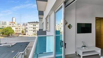 A modern apartment with a balcony overlooking a cityscape, a flat-screen TV mounted on the wall, and a small white side table with a plant.