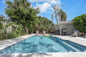 Una piscina rectangular con agua azul cristalina, rodeada de exuberante follaje y palmeras. Se observan dos sillas de descanso y una parrilla de barbacoa al fondo.