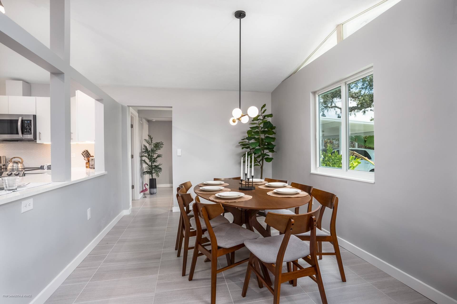 A modern dining area with a round table set for six, surrounded by wooden chairs with grey cushions. A tall, black pendant light hangs above the table.