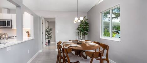 A modern dining area with a round table set for six, surrounded by wooden chairs with grey cushions. A tall, black pendant light hangs above the table.