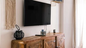 Una TV a schermo piatto montata a parete, un mobiletto in legno con cassetti, un arazzo macramè e uno scaffale con libri e oggetti decorativi.