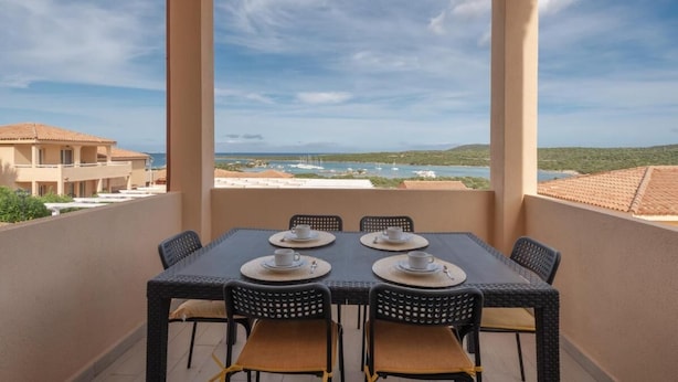 A balcony with a table set for two, overlooking a coastal landscape with buildings and boats. - Stunning Views of Baia de Bahas Residence 1 Bedroom Sleeps 4 (Golfo Aranci)