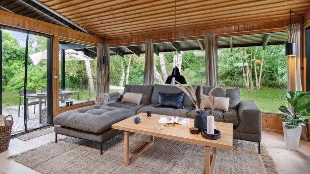 A modern living room with a wooden ceiling, large windows, a grey sectional sofa, a wooden coffee table, and a view of a lush garden.