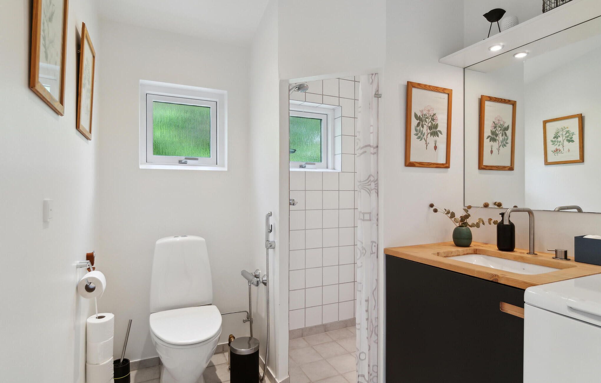 A modern bathroom with a toilet, shower, and sink. There are framed botanical prints on the wall and a window with a view of greenery.