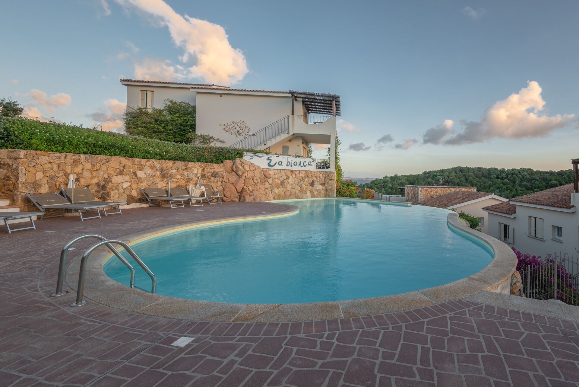 A swimming pool with lounge chairs, a stone wall, and a building with a balcony.
