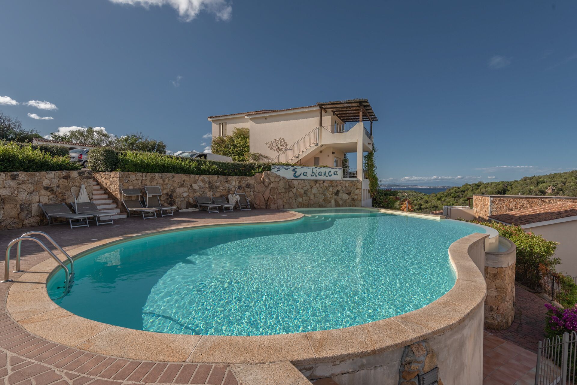 A swimming pool with lounge chairs, a stone wall, and a building with a sign reading "Eabiana".