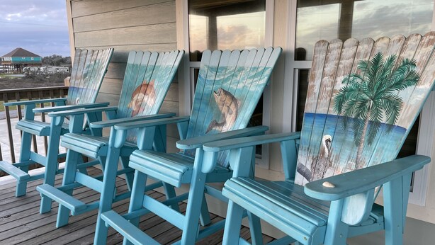 Une rangée de chaises Adirondack turquoise, dont les dossiers sont décorés de motifs de poissons peints et de palmiers, est disposée sur une terrasse en bois.