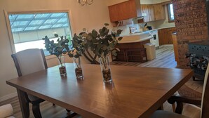 Un comedor con una mesa de madera, sillas y tres jarrones con plantas. Se observa una araña colgante sobre la mesa, y una ventana con persianas a la izquierda.