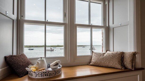 Un ventanal con vistas a barcos en el agua, una preparación para el desayuno con una tetera y tazas, y cojines decorativos.