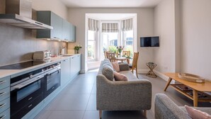 A modern kitchen with grey cabinets, a built-in oven, and a dining area with a grey sofa and wooden table. - Trehafod, Aldeburgh (Aldeburgh)
