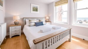 A bedroom with a wooden bed frame, white bedding, two bedside tables with lamps, a framed picture on the wall, and a view of the sea through large windows. - Trehafod, Aldeburgh (Aldeburgh)