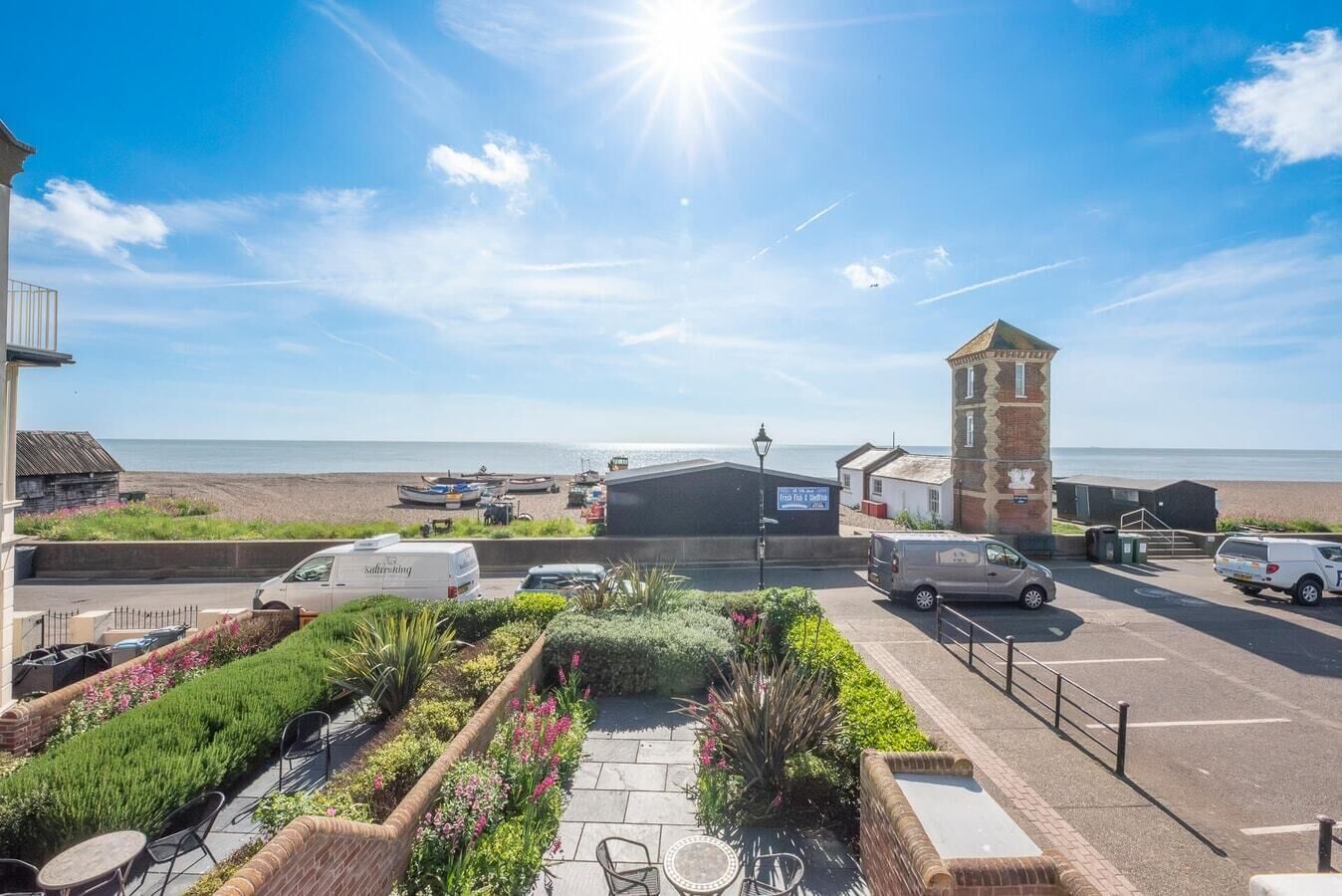 Een kusthotel met een parkeerplaats, een tuin vol planten en bloemen, en uitzicht op het strand en de zee.