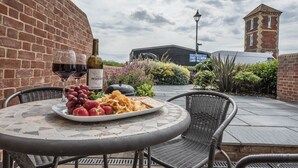 Une terrasse extérieure avec une table ronde, des chaises en osier, une bouteille de vin, des raisins, des fraises et des chips.