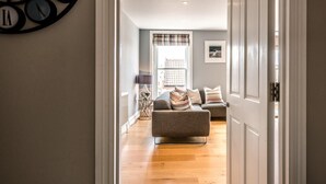 A hallway leading to a living room with a sofa, a clock on the wall, and a window with blinds. - Lyndhurst, Aldeburgh (Aldeburgh)