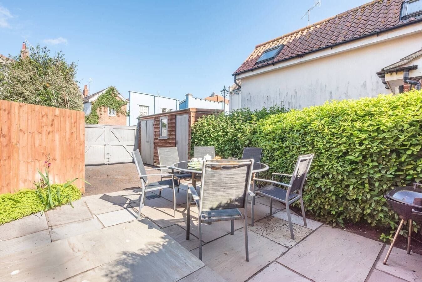 A patio area with a round table and four chairs, a wooden fence, and a hedge.