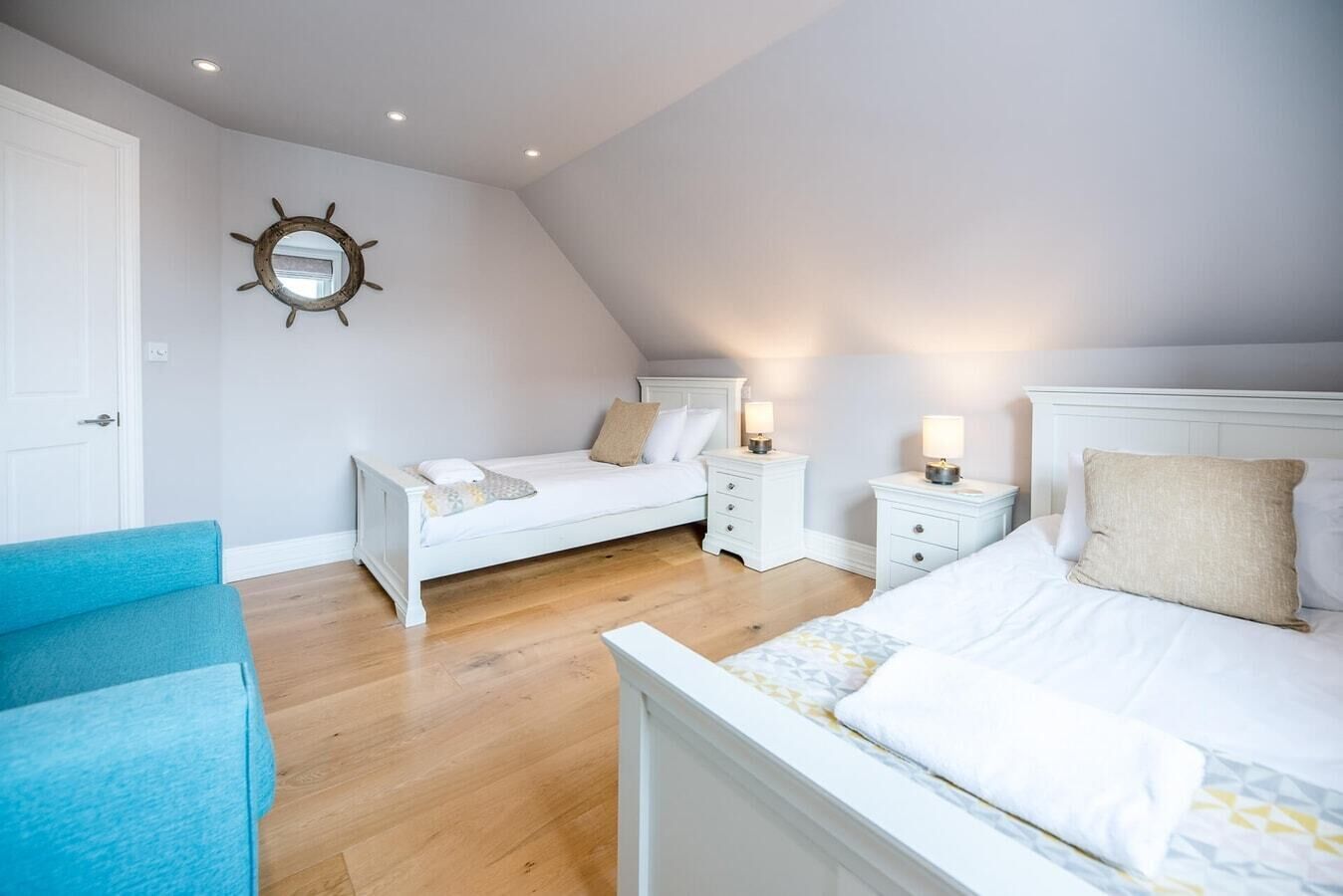 A bedroom with two beds, a blue chair, white nightstands, and a wall-mounted mirror.