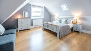 A bedroom with a sloped ceiling, a bed with a headboard, a bedside table with a lamp, a dresser, and a window with a view. - Lyndhurst, Aldeburgh (Aldeburgh)