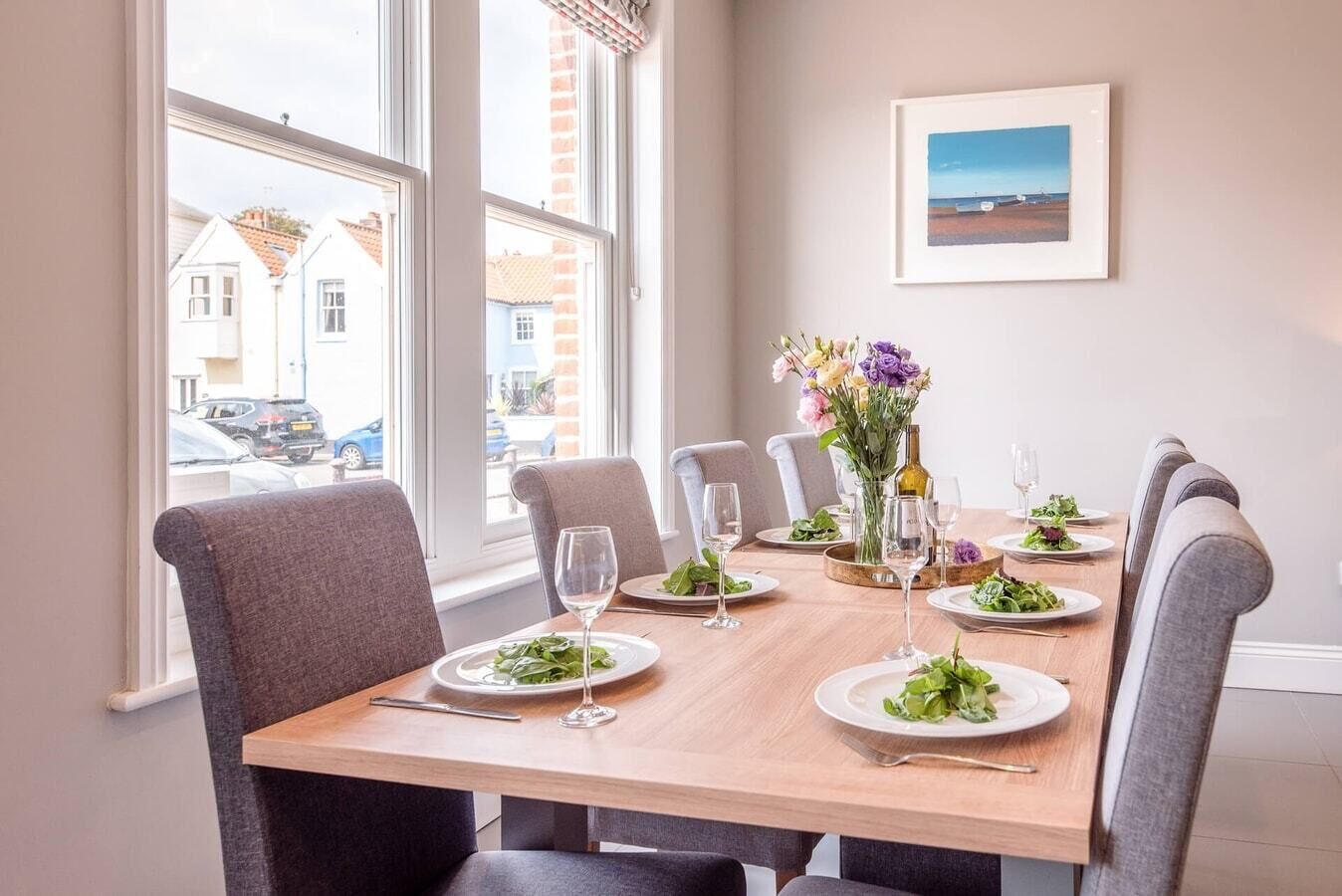 A dining area with a wooden table set for six, six grey upholstered chairs, a vase of flowers, and a framed picture on the wall.