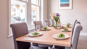 A dining area with a wooden table set for six, six grey upholstered chairs, a vase of flowers, and a framed picture on the wall. - Lyndhurst, Aldeburgh (Aldeburgh)