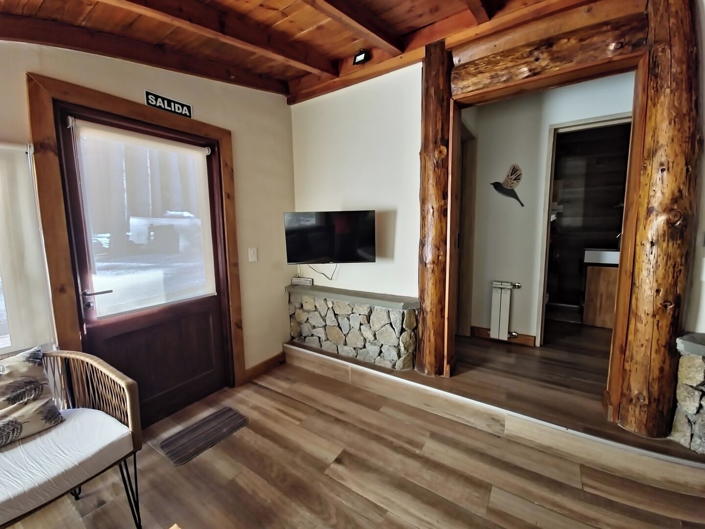 A room with a stone fireplace, wooden beams, a TV mounted on the wall, and a door labeled "SALIDA".