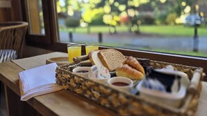 Ein Frühstücksbrett mit Brot, Croissants und Saft auf einem Holztisch neben einem Fenster mit Blick auf Bäume.