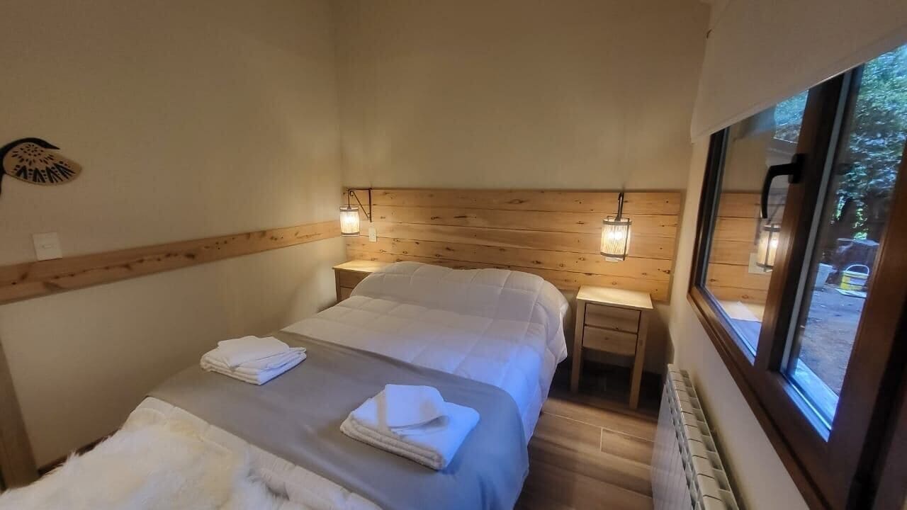 Quarto com cabeceira de madeira, duas mesas de cabeceira com luminárias, cama com roupa de cama branca e vista pela janela.
