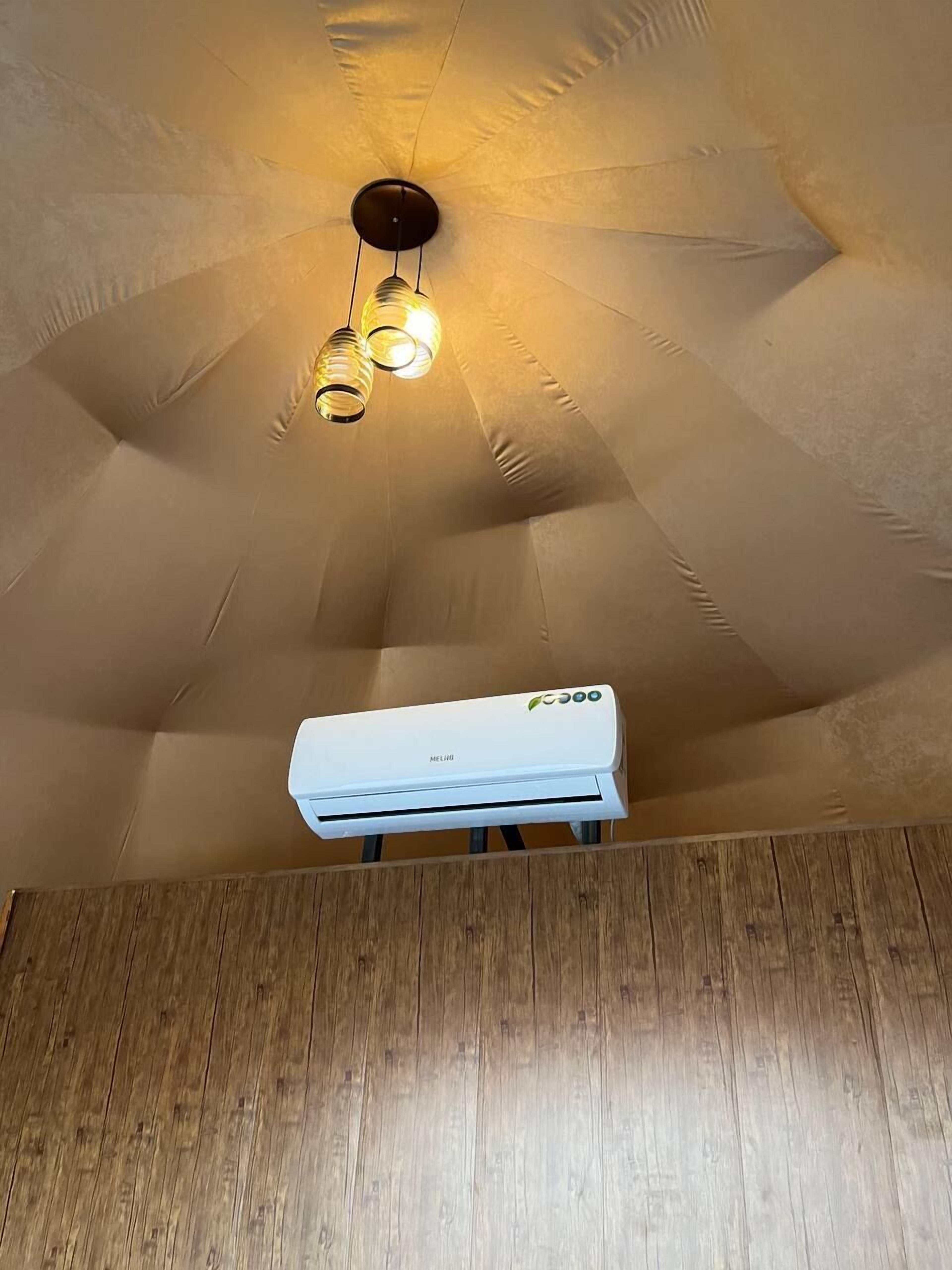 A room with a wooden floor, a white air conditioning unit mounted on the wall, and a ceiling with a unique design and hanging light fixtures.
