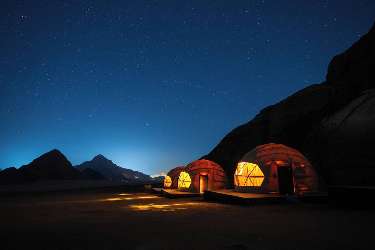 Three dome-shaped tents with warm lighting, set against a backdrop of mountains and a starry night sky.