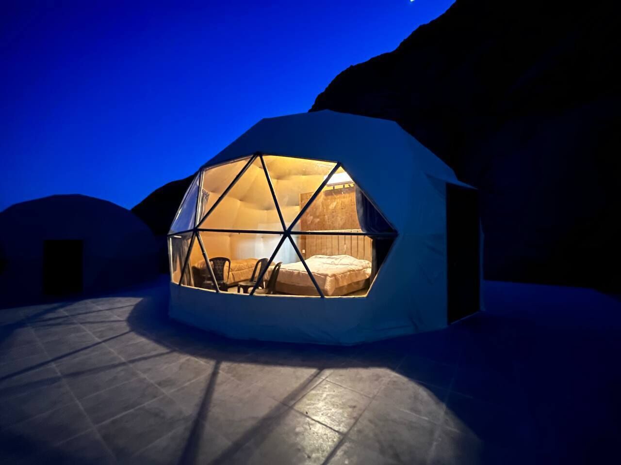 Una estructura de domo geodésico con una cama y zona de estar en su interior, rodeada de un suelo de piedra y otras estructuras similares.
