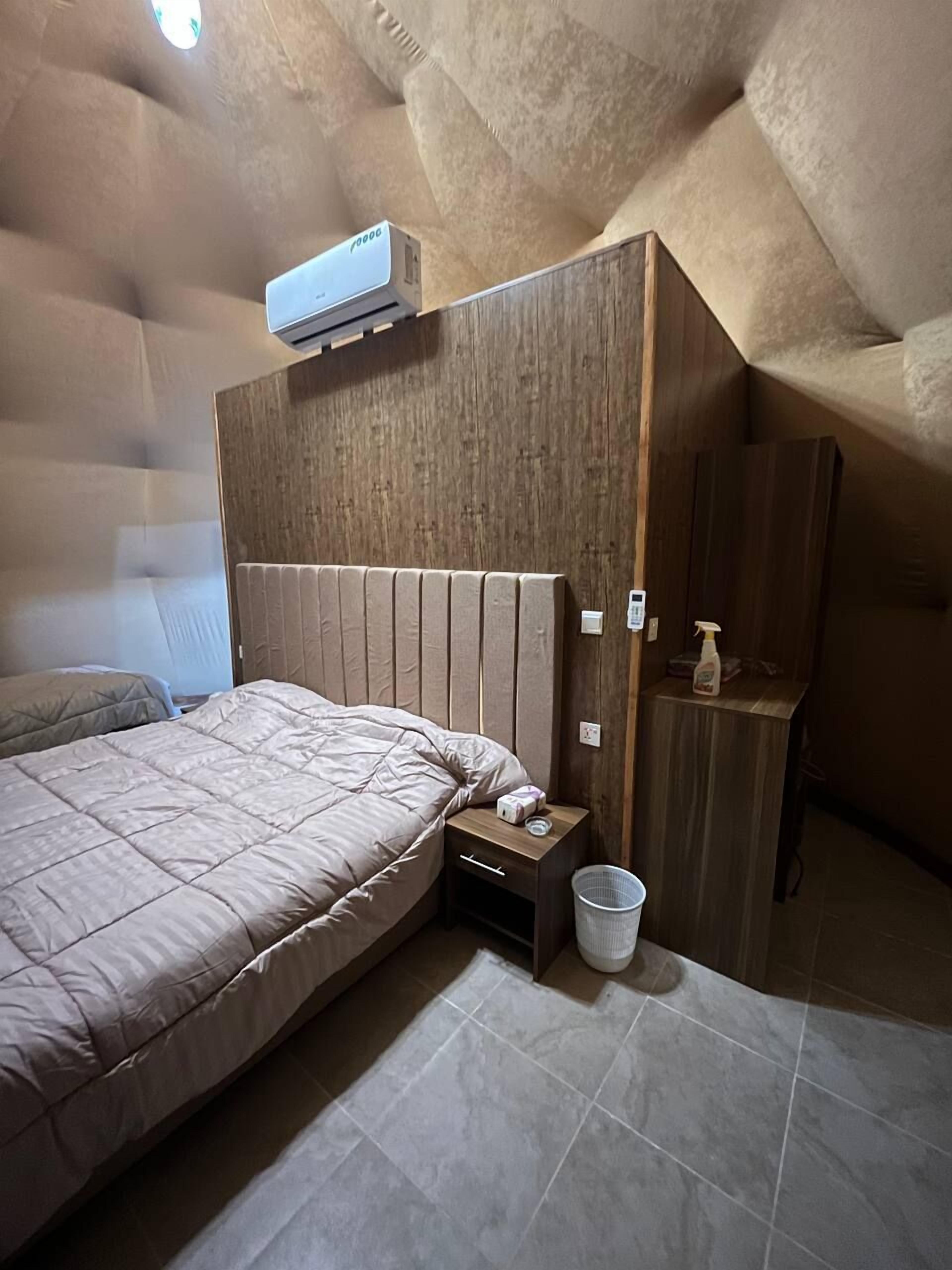 Una camera da letto con un letto grande, una testiera in legno, un comodino, un cestino e un condizionatore d'aria.