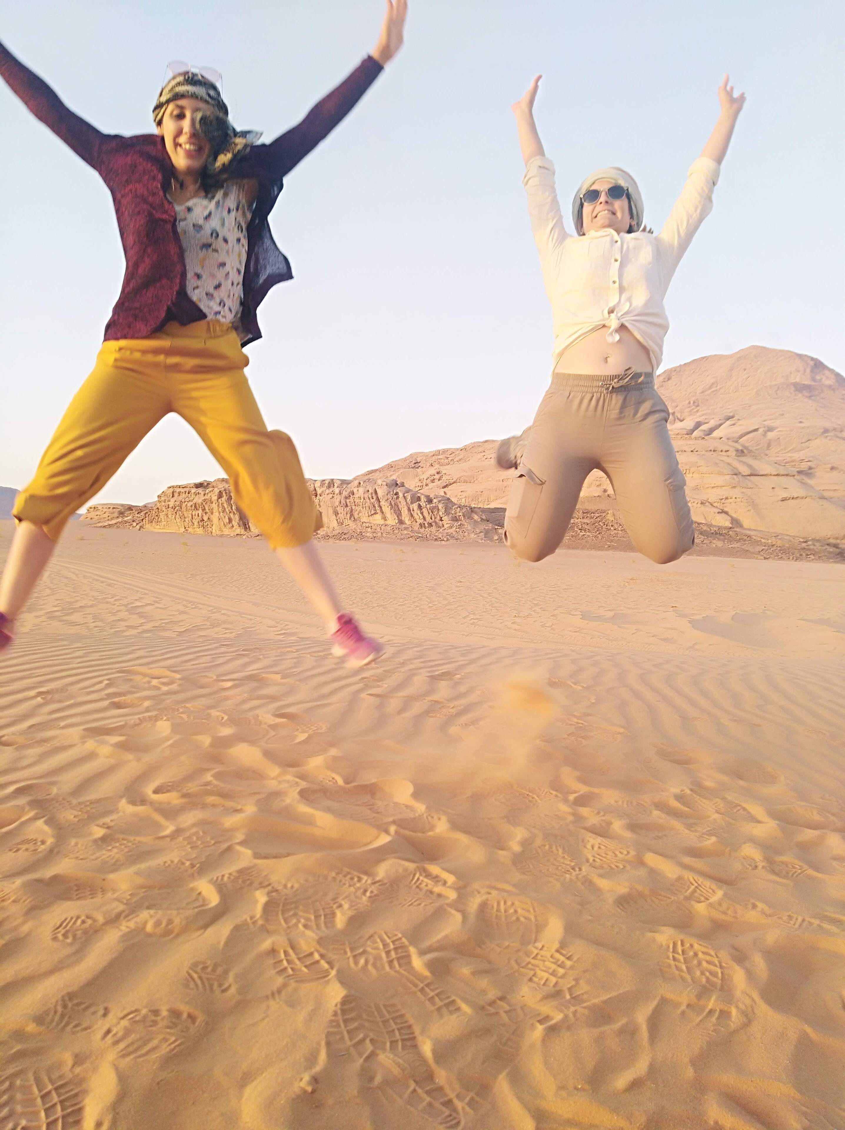 Two people jumping in a desert with footprints in the sand.