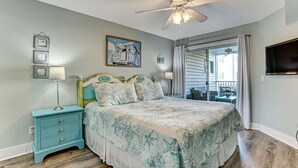 A bedroom with a bed, a nightstand, a ceiling fan, a window with curtains, and a wall-mounted TV. - "Spritz By The Sea” is your beach side paradise! (Tybee Island)
