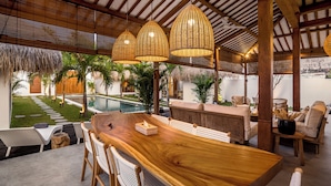 Une table à manger en bois, des chaises en osier, un toit de chaume et une piscine en arrière-plan.