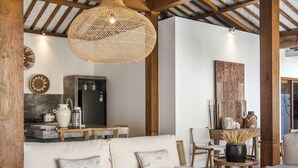 Un salon accueillant avec un canapé, des fauteuils en osier et des petites tables rondes en bois. Un luminaire tressé est suspendu au plafond, et on aperçoit un coin cuisine à l’arrière-plan.