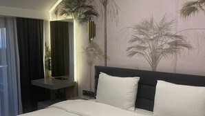 Quarto de hotel com uma cama grande, escrivaninha, espelho e um mural na parede com palmeiras.