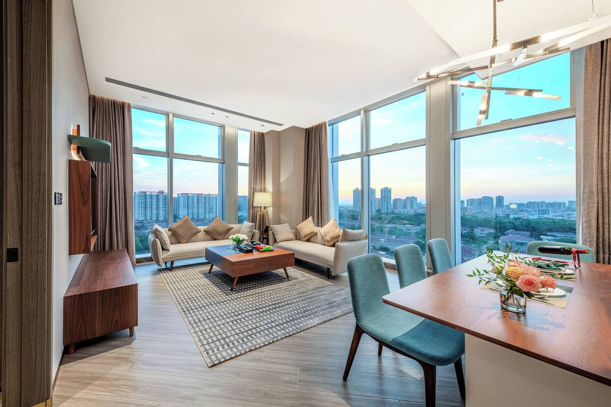 A modern living room with a sofa, coffee table, and dining area, large windows with a city view, and a wooden accent wall.