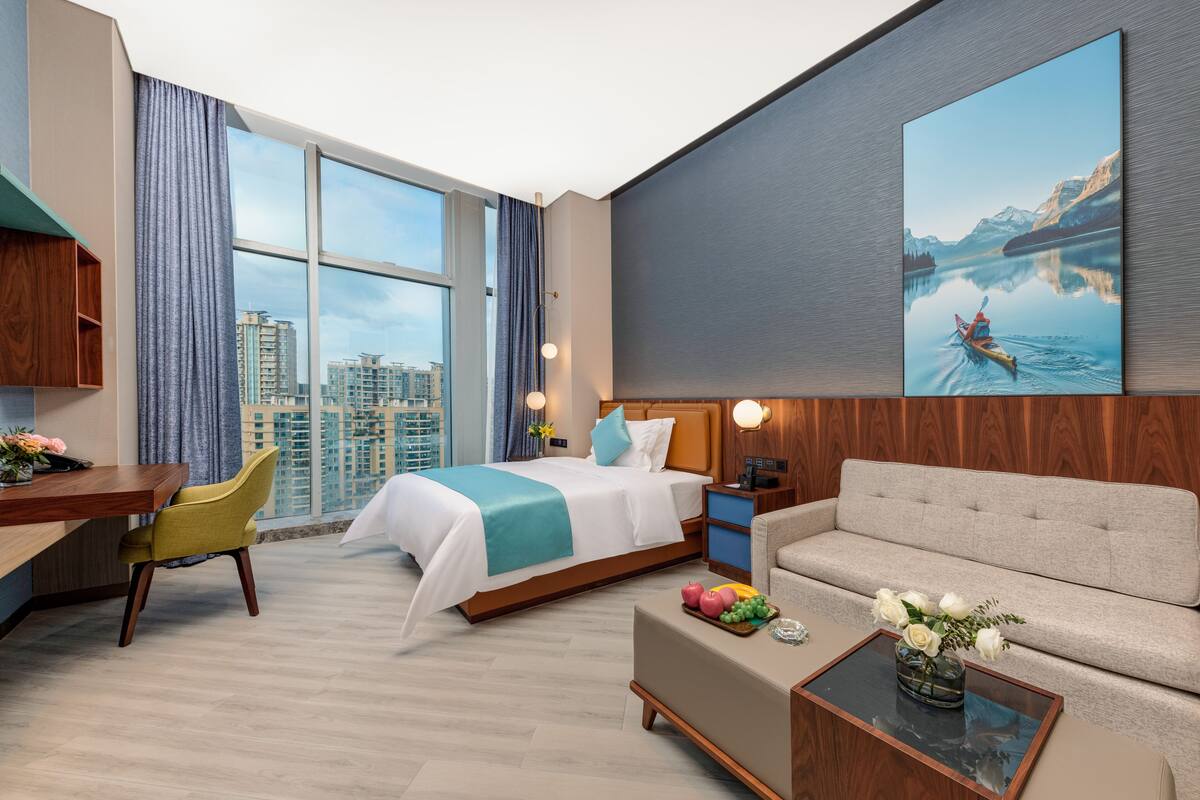A modern hotel room with a large bed, a sofa, a desk, and a view of the cityscape through large windows.