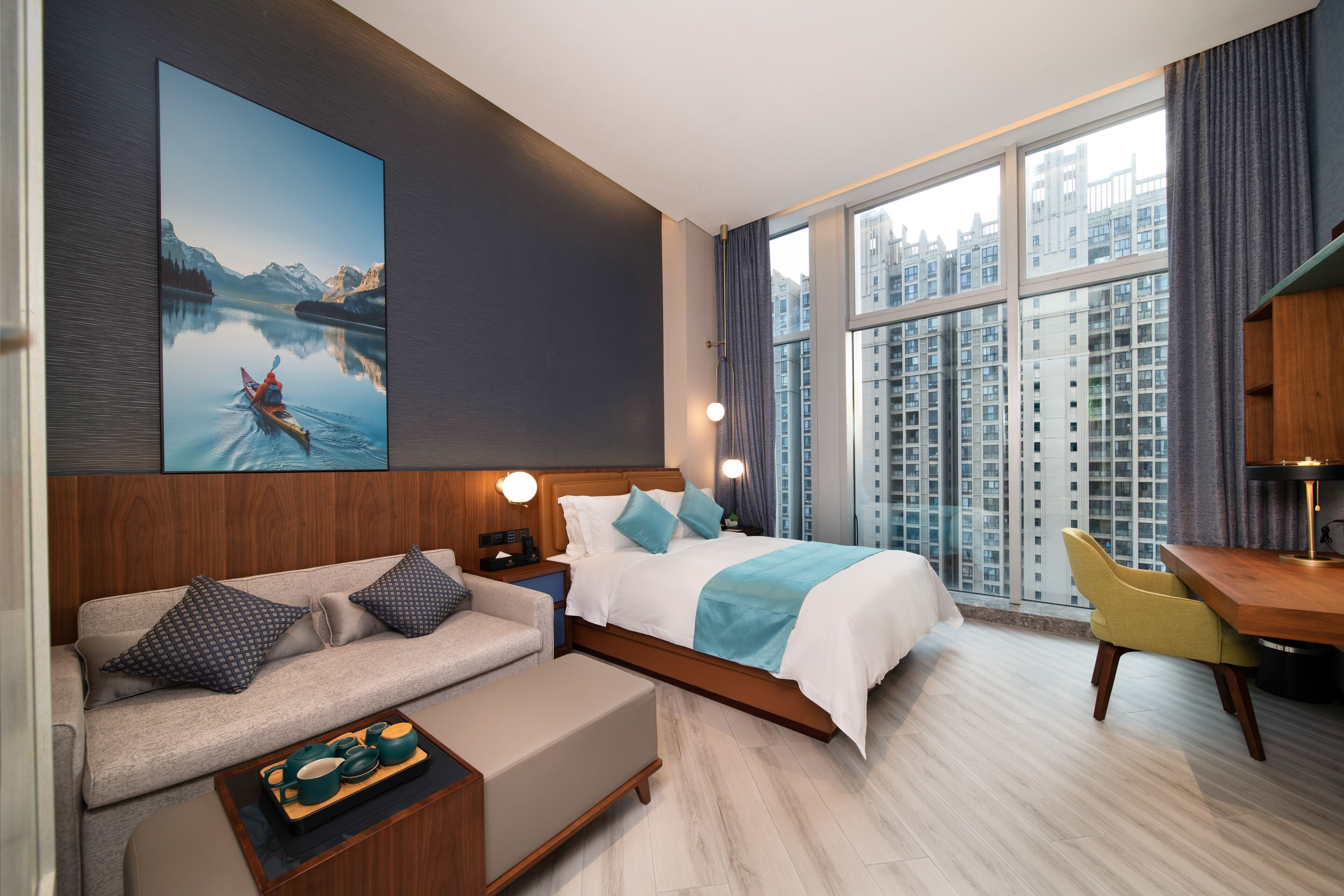 A modern hotel room with a large bed, a sofa, a desk, and a view of high-rise buildings.