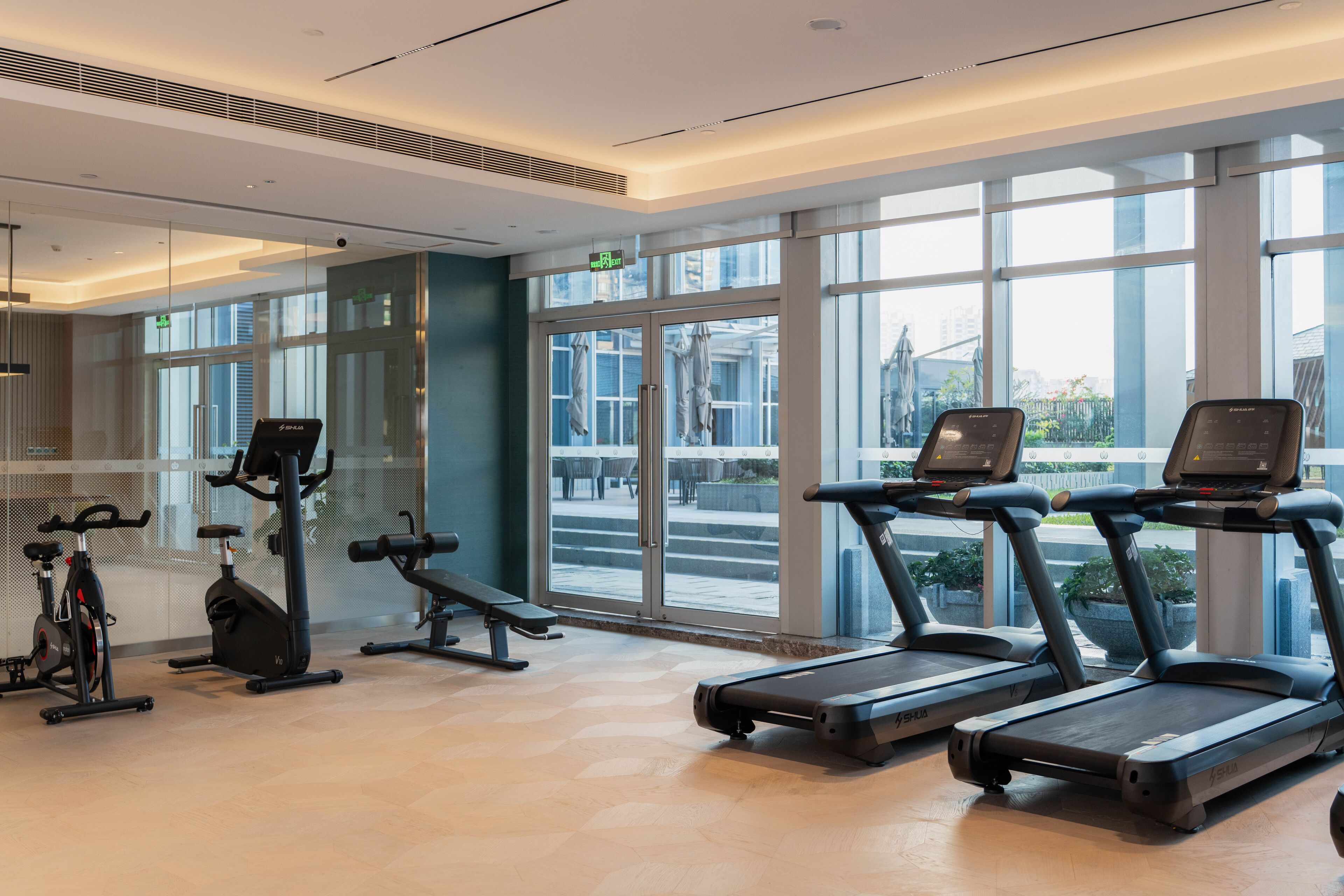A modern gym with treadmills, stationary bikes, and weight machines. Large windows provide natural light and views of the city.
