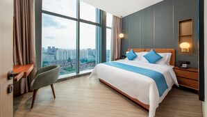 Ein modernes Hotelzimmer mit großem Fenster, einem Bett mit blauer Bettwäsche, einem Sessel, einem Nachttisch und einem Beistelltisch mit Lampe.