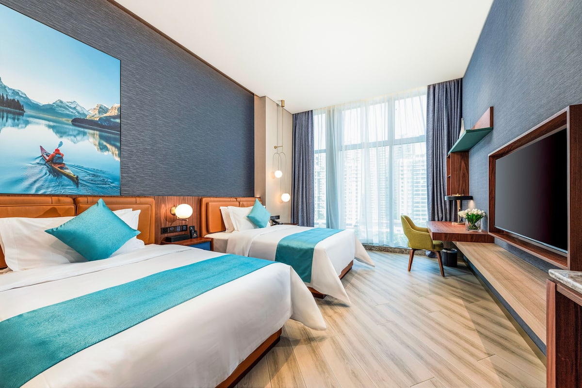 A hotel room with two beds, a desk, a TV, and a large window with curtains.