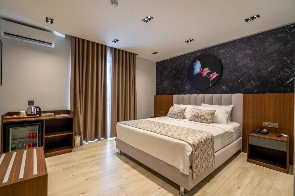 A modern hotel room with a large bed, a desk with a kettle, a small side table, and a wall-mounted air conditioner.