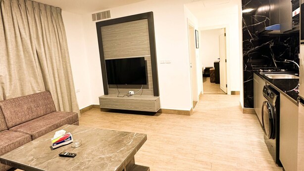 A modern living room with a flat-screen TV, a sofa, a coffee table, and a kitchen area with a stove and washing machine. - Central Apartment For Rent 30 (Amman)