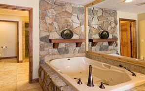 A bathroom with a stone wall, a large bathtub, and a mirror above it.