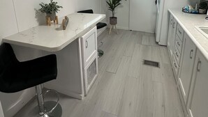 A modern kitchen with white cabinetry, a marble countertop, a black bar stool, a potted plant, and a window with blinds. - Luxury Camping at Calusa (Key Largo)