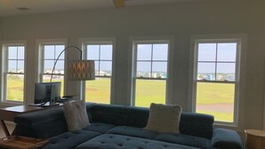 A living room with a sectional sofa, a ceiling fan, and large windows offering a view of a green field. - Sun-kissed Mermaid / 846 Ocean Blvd West 2 Bedroom Home by RedAwning (Supply)