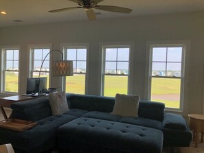 A living room with a sectional sofa, a ceiling fan, and large windows offering a view of a green field. - Sun-kissed Mermaid / 846 Ocean Blvd West 2 Bedroom Home by RedAwning (Supply)