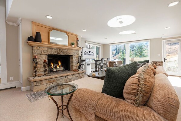 A living room with a stone fireplace, a glass coffee table, and a beige sofa with green cushions. - Two-level corner condo w/ fireplace, amazing mountain views, spa and hot tub access (Vail)