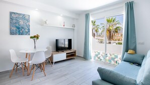 A modern living room with a sofa, a TV, a dining table with chairs, and a balcony with a view of palm trees and buildings. - 475 B Island Village Sea View Pool & Terrace (Costa Adeje)