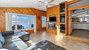 A spacious living area with a large window, a ceiling fan, a flat-screen TV, and a kitchen area with cabinets and a countertop.